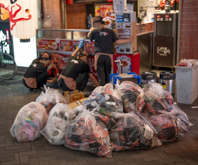 A narrow urban alleyway cluttered with discarded rubbish and debris, with a large wheeled garbage container positioned centrally in the foreground. The container appears to be made of a durable, weath