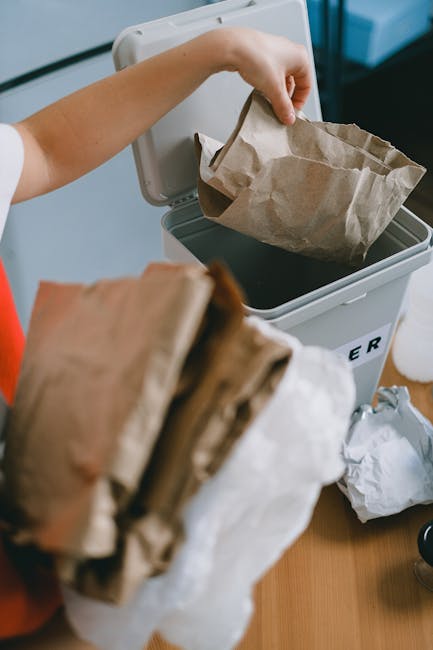 A person is seen disposing of crumpled brown paper bags into a white bin labeled with black text, possibly on a wooden surface. One hand is holding an opened paper bag above the bin, which is partiall
