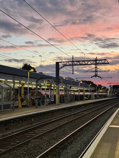 The image depicts a railway station platform during dusk, with the sky painted in shades of pink, orange, and purple, indicating a sunset. In the foreground, several train tracks run parallel, with ev