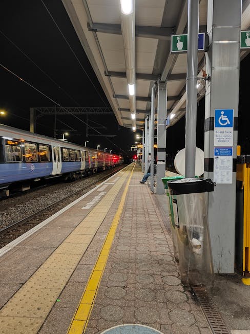 The image depicts a railway station platform during dusk, with the sky painted in shades of pink, orange, and purple, indicating a sunset. In the foreground, several train tracks run parallel, with ev