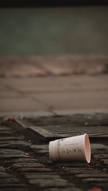 A black, cylindrical public rubbish bin with a textured, weathered surface stands on a cobblestone street, overflowing with various discarded items. The lid of the bin is partially open, supporting se